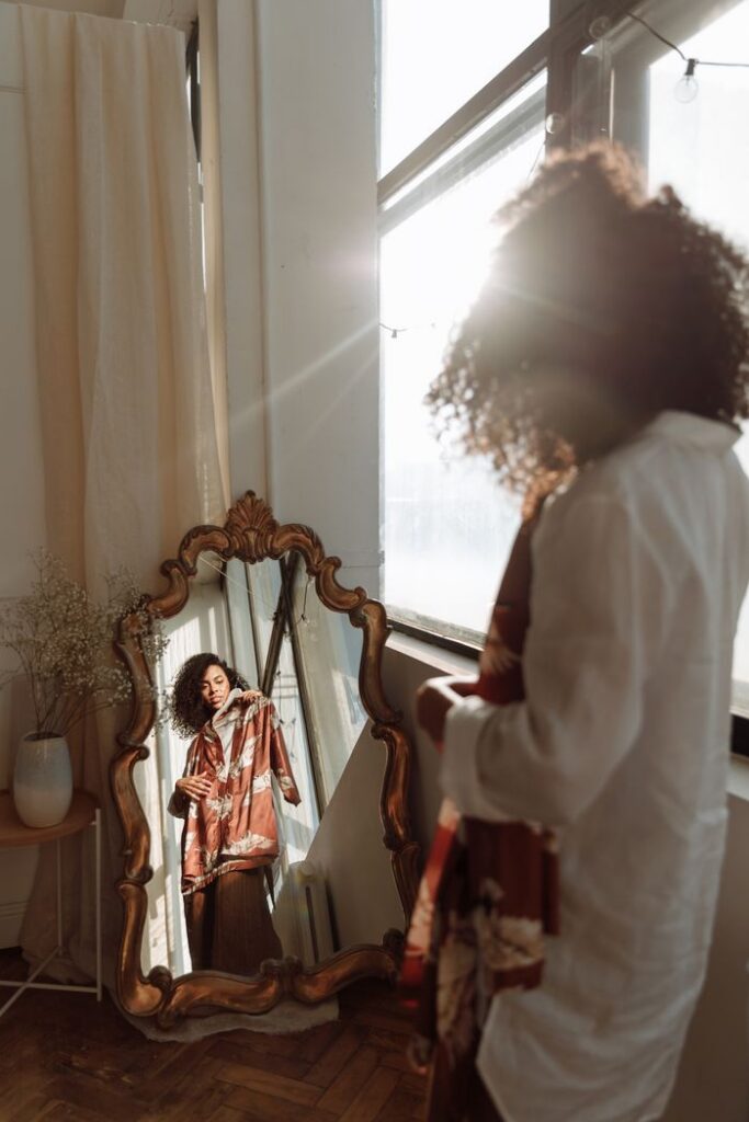 Elegant woman in reflection, holding clothes in sunny room.